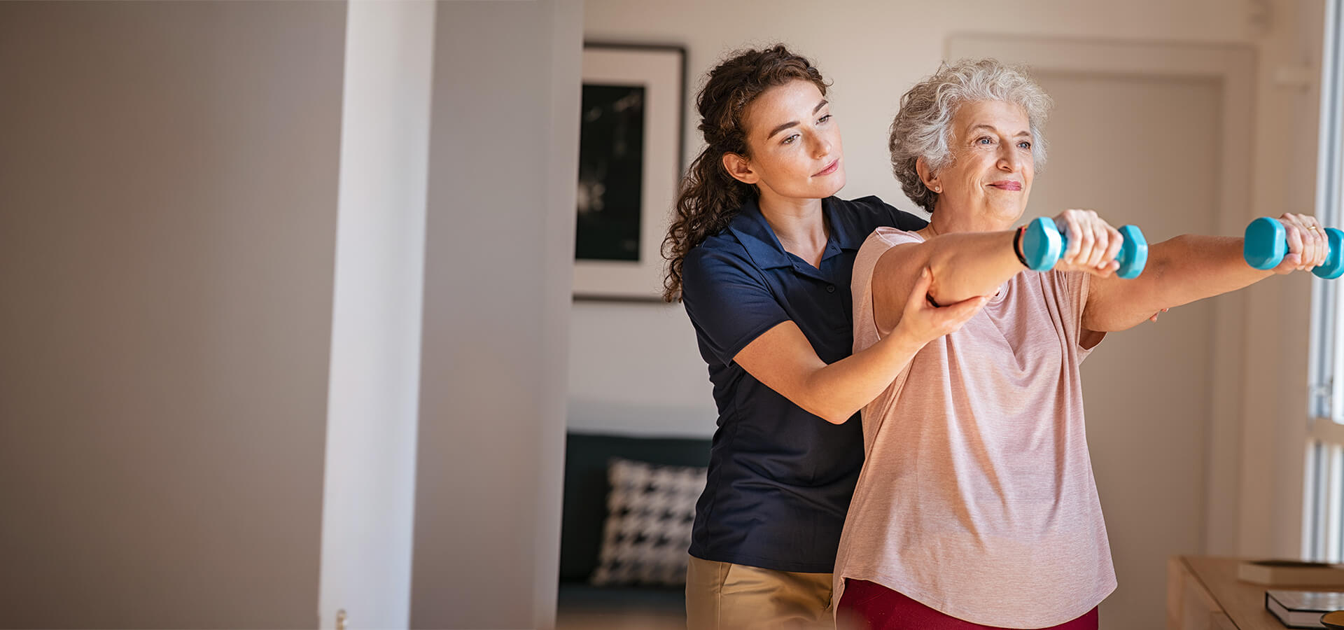 Caregiver assists senior woman with light exercise using dumbbells indoors.