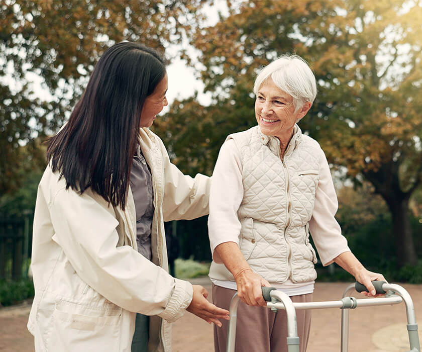 Elderly woman with walker being assisted by a caregiver outdoors in an autumn setting.