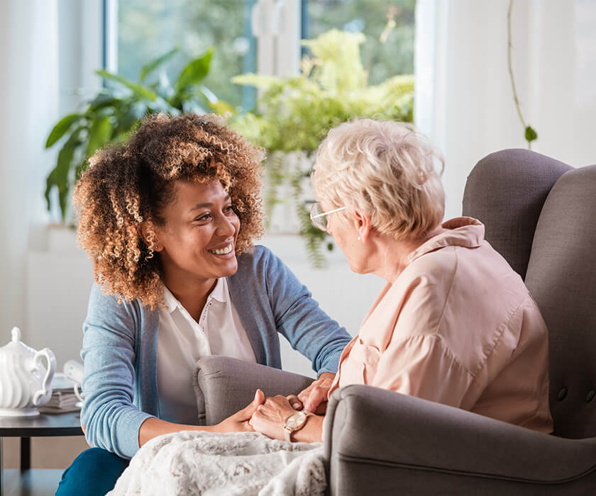 Caregiver smiling and supporting an elderly woman in a cozy living room.