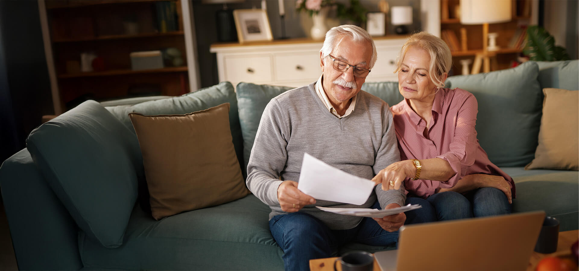 Elderly couple reviewing documents together on a sofa next to a laptop.