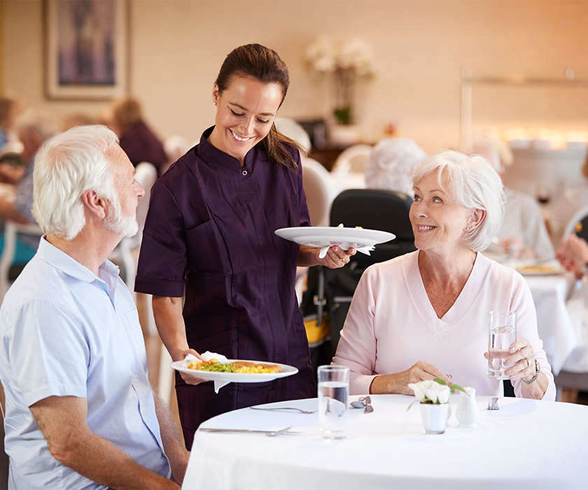 Friendly server delivering meals to two seniors in a dining hall setting.