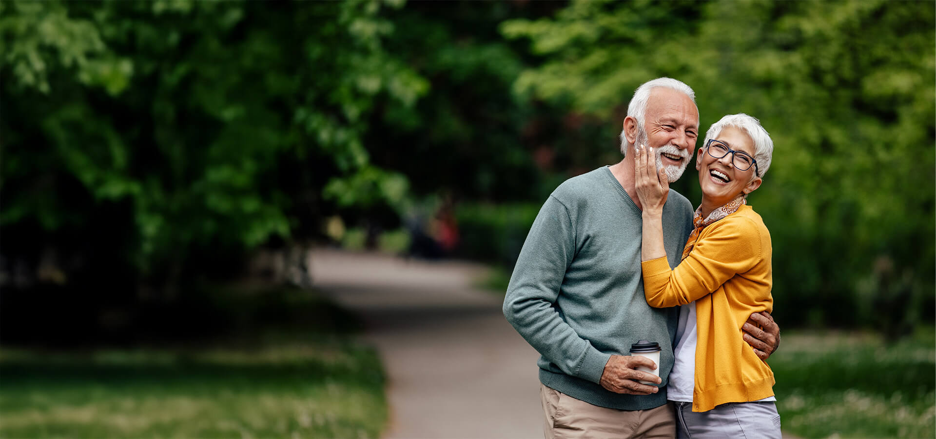 Elderly couple smiling while embracing in a lush, green park.