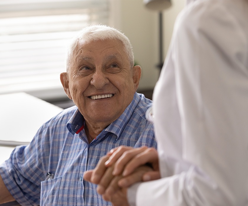 Smiling elderly man holds a caregivers hand in a cozy living space.