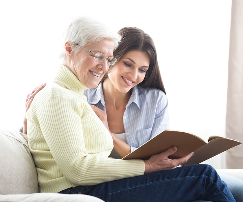 Elderly woman reading a book with a younger woman on a couch in a bright room.
