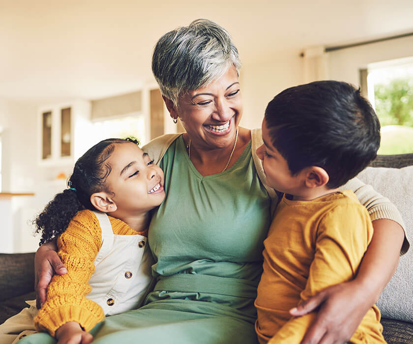 Smiling elderly woman hugging two children on a couch in a bright living room.