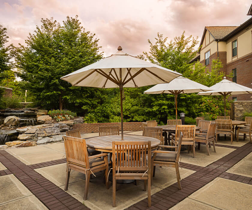 Outdoor dining area with wooden tables and chairs under umbrellas beside lush greenery.