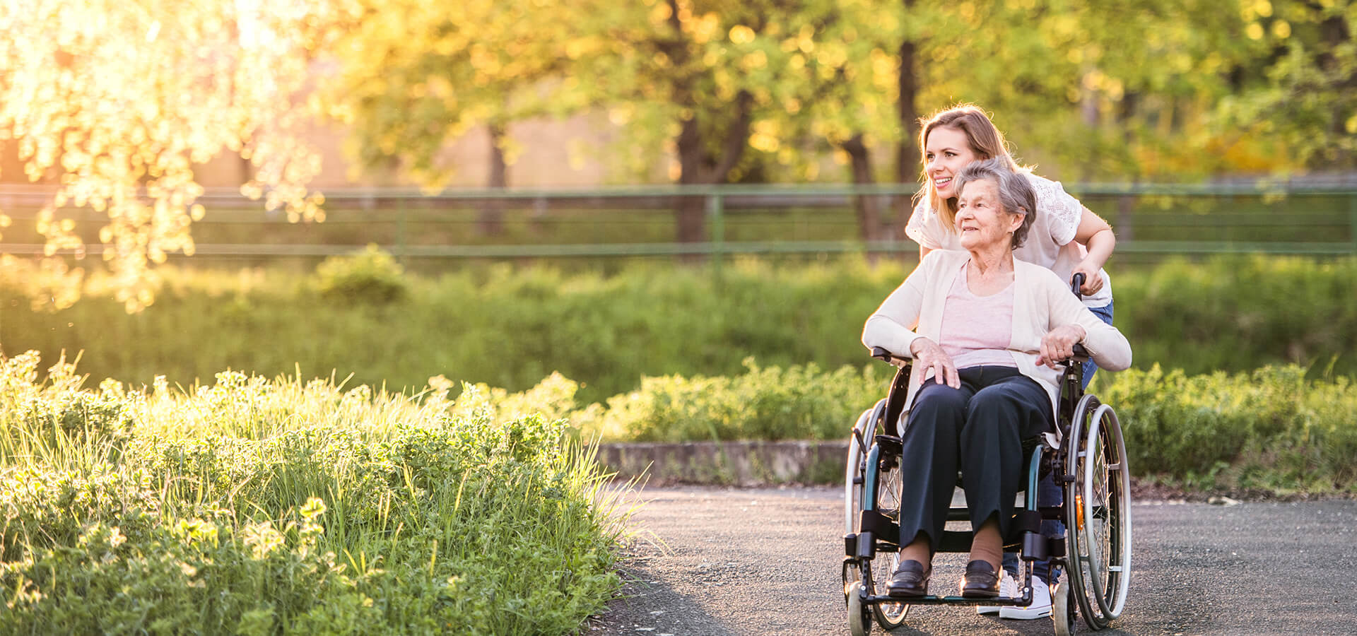 Caregiver pushing an elderly woman in a wheelchair outdoors on a sunny day.