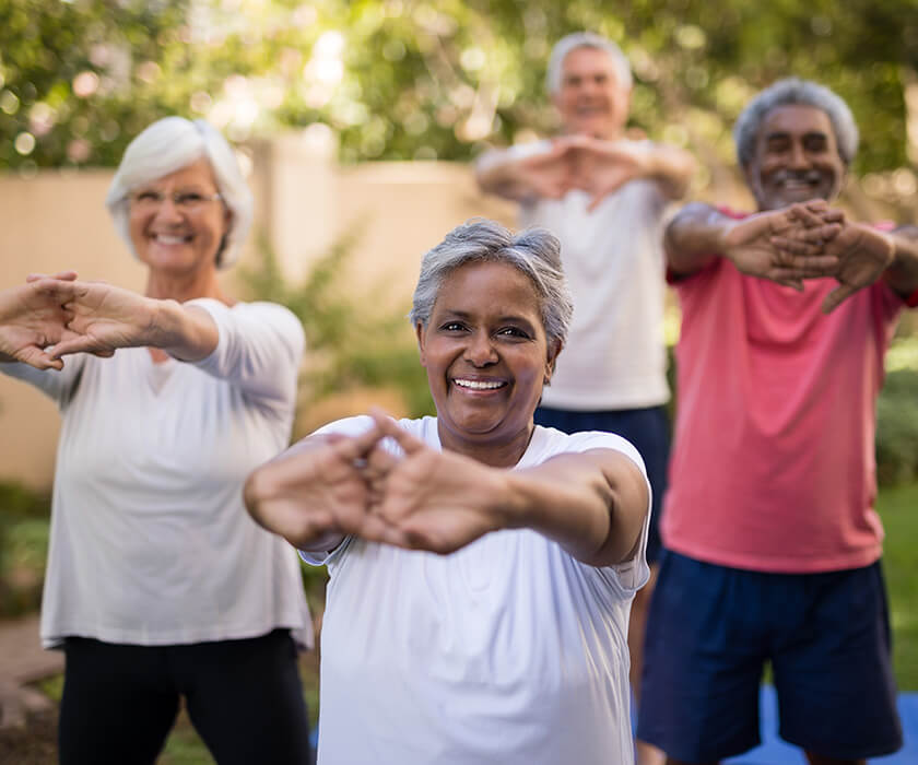 Seniors practicing yoga outdoors smiling and stretching arms forward.