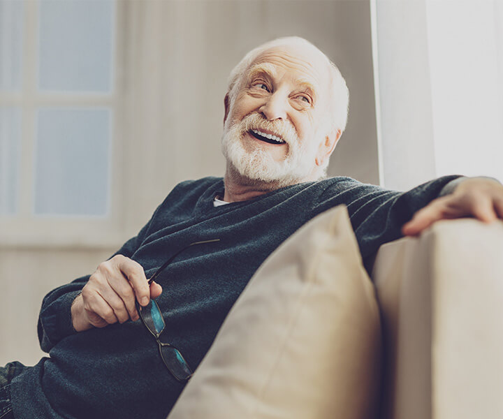 Elderly man smiling while relaxing on a sofa in a sunlit room.