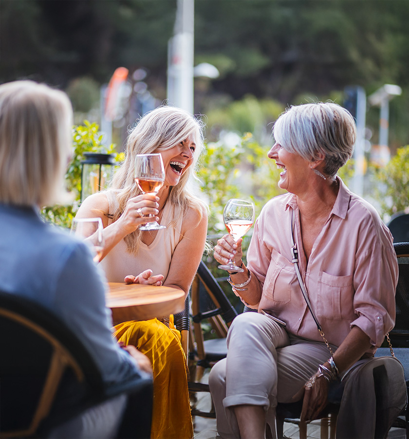 Three women laughing and enjoying drinks at outdoor seating area.