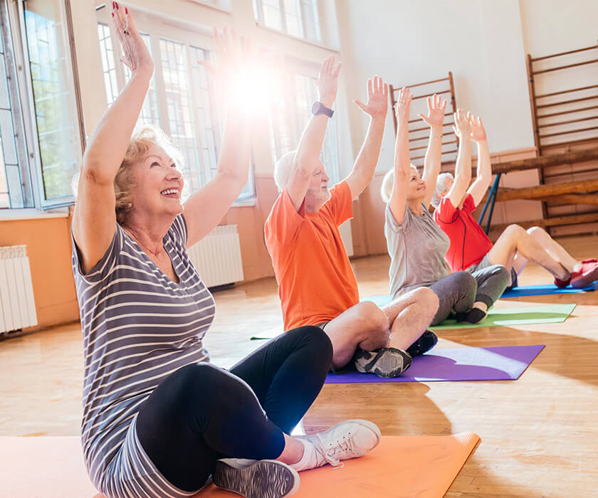 Group of seniors exercising on mats in a sunlit room with raised arms.