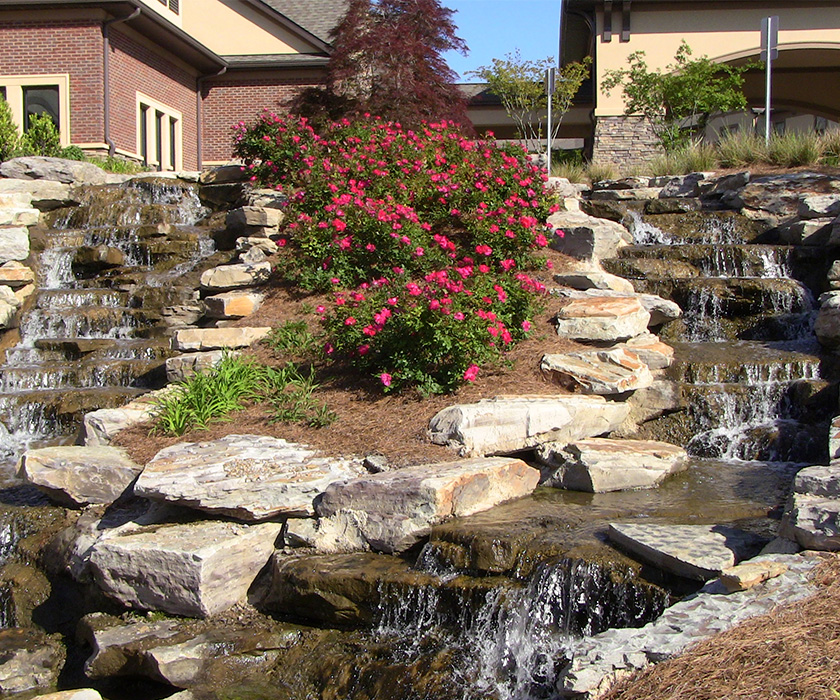 Beautiful rock waterfall and vibrant flowers in front of a brick building.