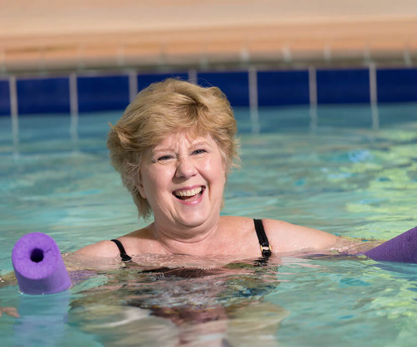 Smiling woman enjoying swimming in a pool with a purple float at a community center.