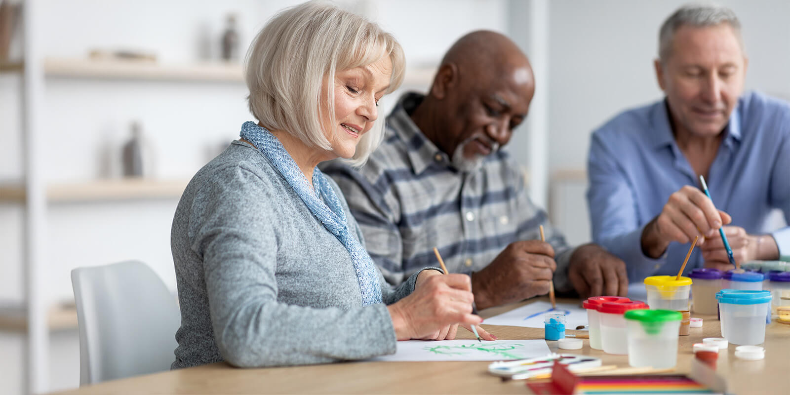 Woman smiling as she and two men paint pictures at a table